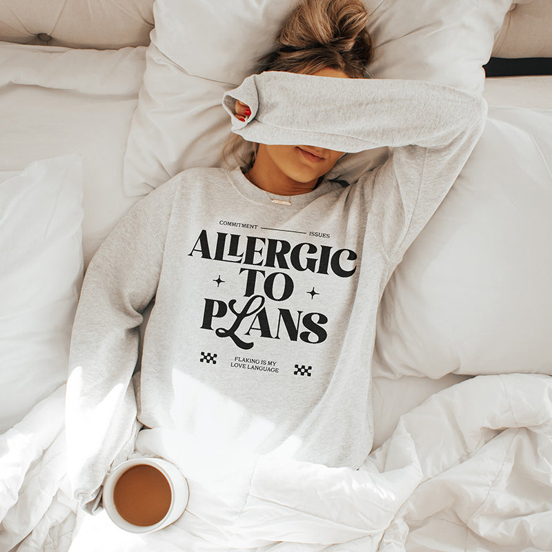 Woman lounging in bed wearing the Allergic to Plans ash grey sweatshirt, holding a coffee mug — cozy introvert aesthetic.