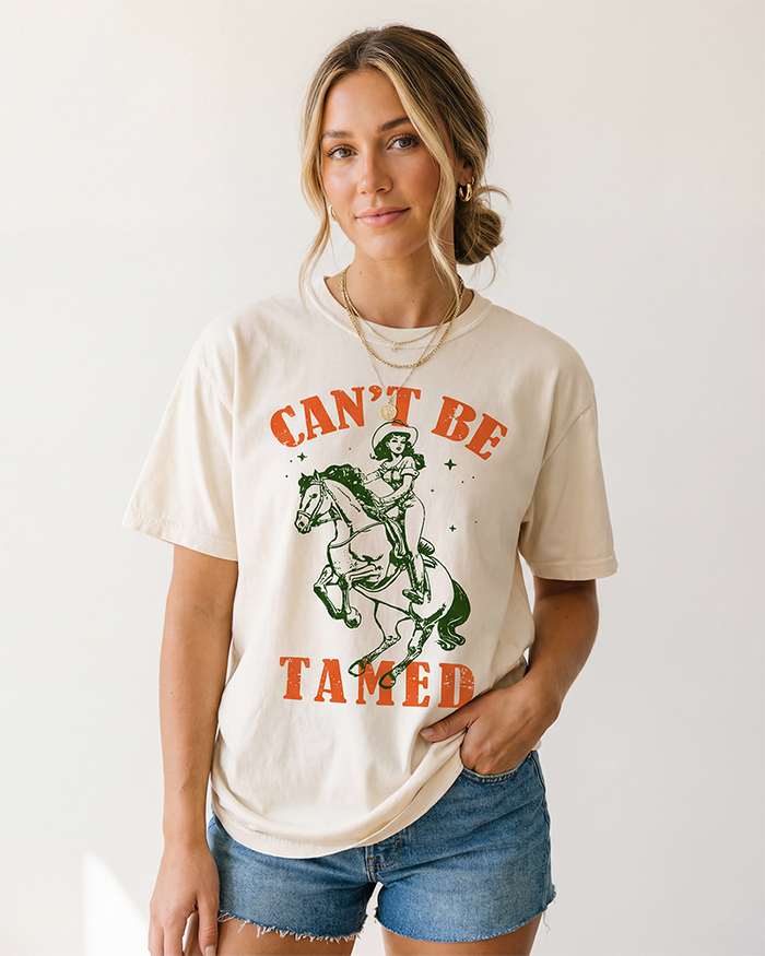 Woman modeling an Ivory Can't Be Tamed Western cowgirl graphic tee featuring vintage horseback rider artwork with denim shorts.