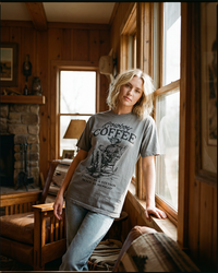 Woman wearing a grey Cowboy Coffee heavyweight graphic tee with vintage Western skeleton cowboy design.