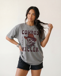 Woman modeling a Grey Cowboy Killer Western skull tee with distressed vintage artwork and black denim shorts.