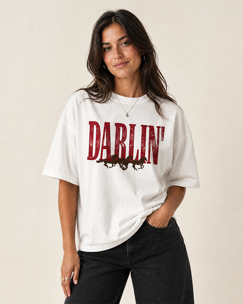 Woman modeling a white heavyweight cowgirl graphic tee featuring Darlin' lettering and running horses.