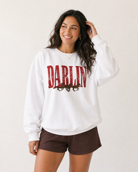 Woman wearing a white Darlin' Western horse sweatshirt with distressed red lettering, brown horse graphic and brown shorts.