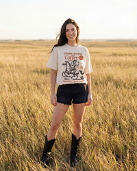 Woman modeling an ivory Drink Some Coffee Western graphic T-shirt with vintage cowgirl and horse artwork.