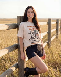 Woman wearing an ivory Western cowgirl graphic tee with horse and coffee design, black shorts and cowboy boots.