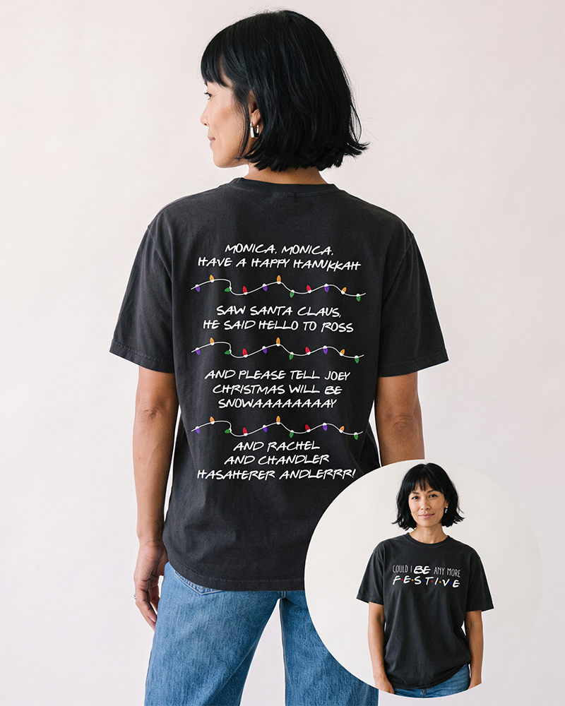 Woman with short black bob wearing a black Friends Christmas heavyweight graphic tee with blue denim jeans, featuring holiday song lyrics on the back and festive lettering on the front.