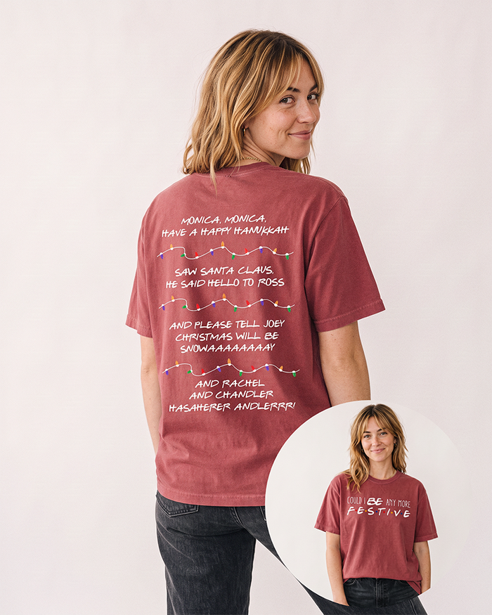 Blonde woman wearing a brick red Could I Be Any More Festive heavyweight tee with vintage black jeans, featuring colorful Christmas lights and Friends-inspired holiday lyrics on the back.