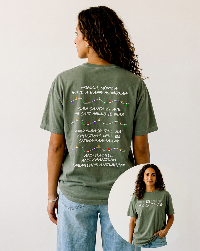 Woman with long curly brunette hair wearing a hemp green Friends-inspired Christmas heavyweight tee with light blue jeans, showing Phoebe's holiday song on the back and festive front graphic.