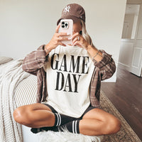Woman wearing ivory Game Day Comfort Colors tee layered with flannel, baseball cap, biker shorts, and socks — cozy sports mom outfit for home or game day.