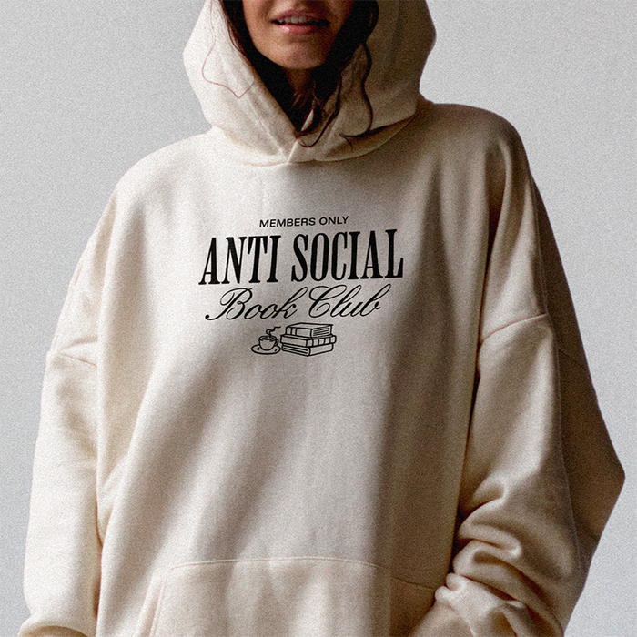 Woman wearing heather dust Antisocial Book Club hoodie while reading a book indoors – cozy book lover hoodie for introverts and homebodies