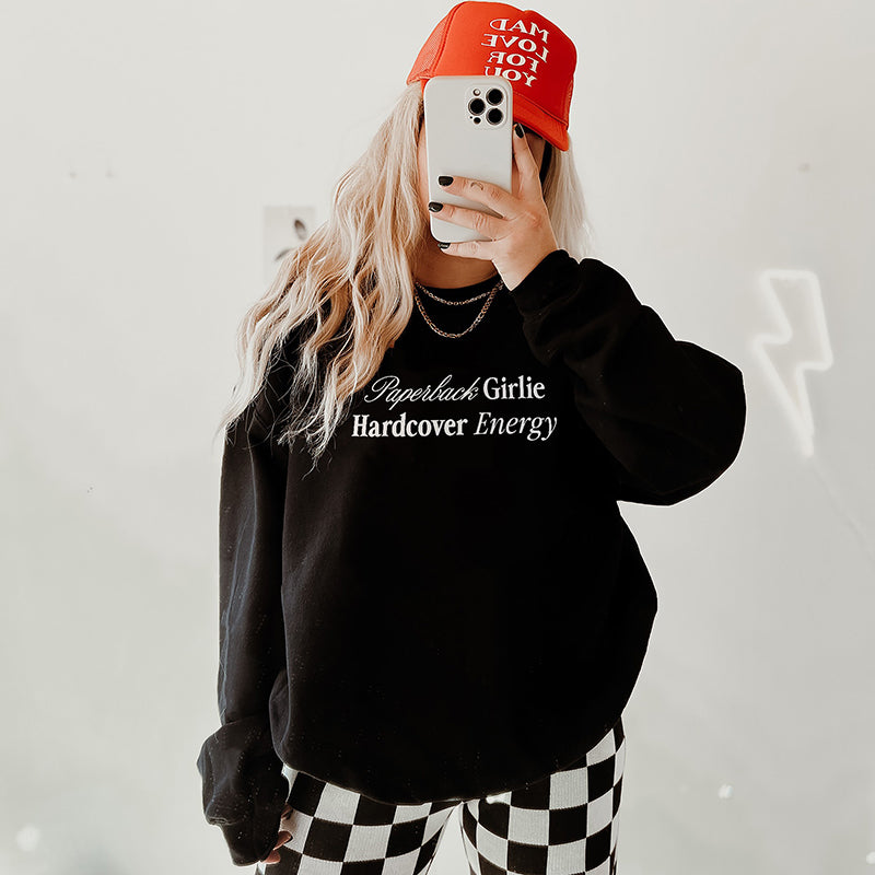Model in black Paperback Girlie Hardcover Energy sweatshirt (Gildan style) styled with a trucker hat and black-and-white checkered pants — casual bookish vibes.