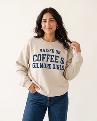 Woman modeling a sand Raised On Coffee & Gilmore Girls crewneck sweatshirt with distressed navy collegiate lettering and blue jeans.