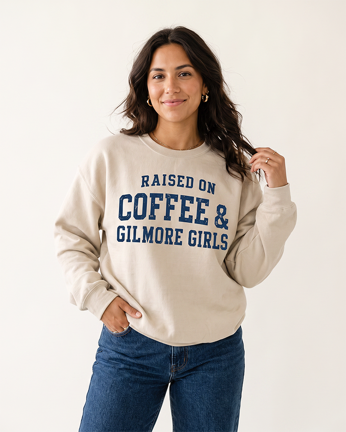 Woman modeling a sand Raised On Coffee & Gilmore Girls crewneck sweatshirt with distressed navy collegiate lettering and blue jeans.