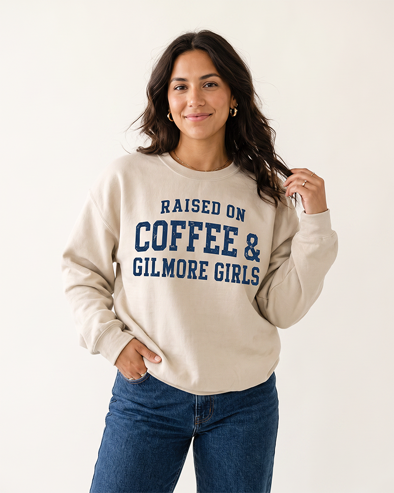 Woman modeling a sand Raised On Coffee & Gilmore Girls crewneck sweatshirt with distressed navy collegiate lettering and blue jeans.