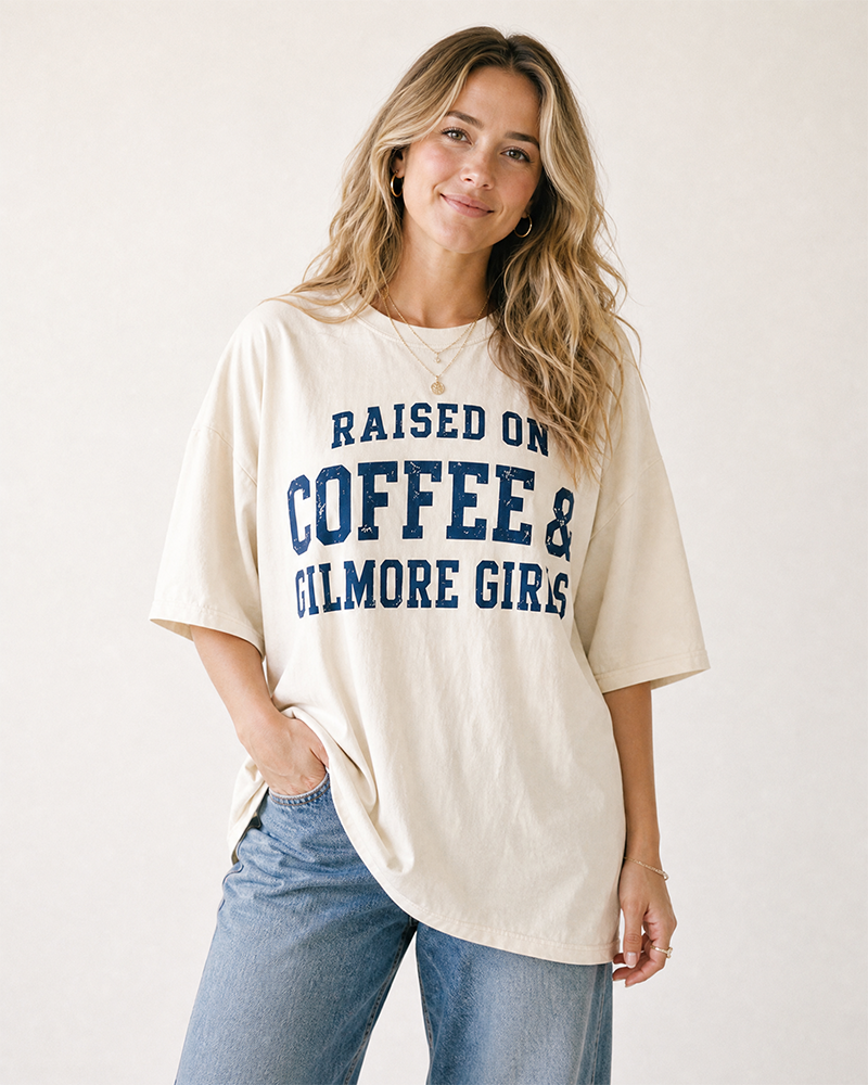 Woman modeling an oversized ivory Gilmore Girls graphic T-shirt with navy Raised On Coffee & Gilmore Girls lettering and blue jeans.