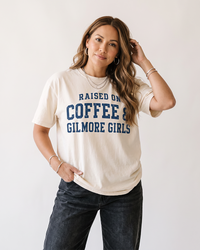 Woman wearing an ivory Raised On Coffee & Gilmore Girls heavyweight graphic tee with distressed navy collegiate lettering.