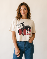 Woman wearing a creme Rodeo Western boxy crop top with distressed cowboy and bucking horse graphic and blue jeans.