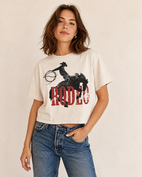Woman wearing a cream Rodeo Western boxy crop top with distressed cowboy and bucking horse graphic and blue jeans.