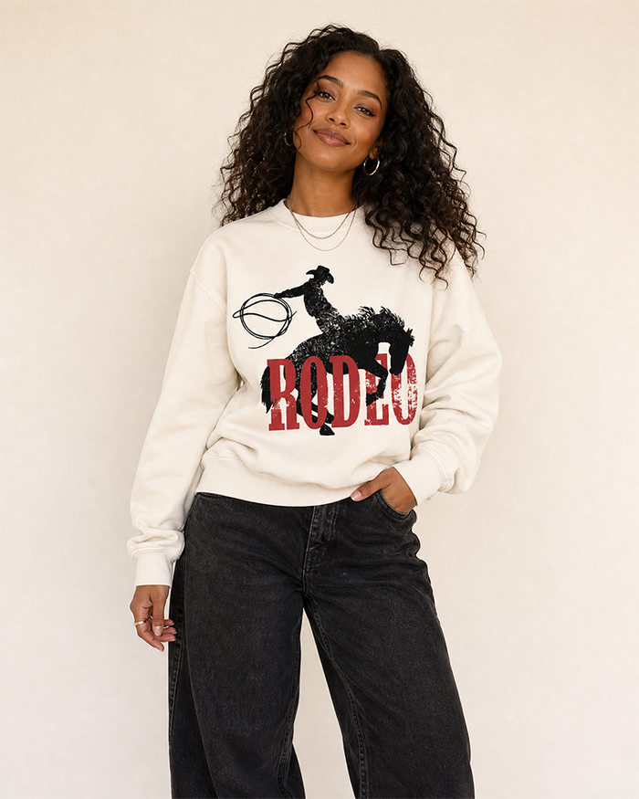 Woman modeling a bone Western rodeo sweatshirt featuring a distressed cowboy, bucking horse and red lettering.