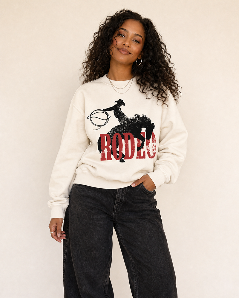 Woman modeling a bone Western rodeo sweatshirt featuring a distressed cowboy, bucking horse and red lettering.
