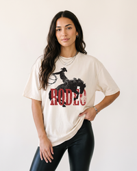 Woman wearing an ivory Rodeo Western heavyweight tee with distressed cowboy and bucking horse graphic and black pants.