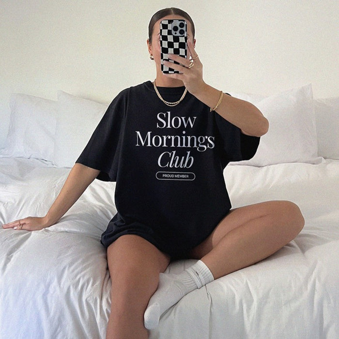 Woman sitting on bed in black Slow Mornings Club Tee, relaxed slow morning aesthetic