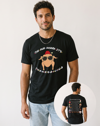 Male model wearing a black Friends-inspired Thanksgiving T-shirt with blue jeans, featuring a turkey in sunglasses on the front and Thanksgiving episode artwork on the back.
