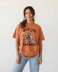 Woman wearing a Yam Wild Like The West Western graphic tee with cowboy and bucking horse artwork and light wash jeans.