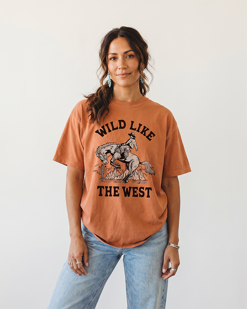 Woman wearing a Yam Wild Like The West Western graphic tee with cowboy and bucking horse artwork and light wash jeans.