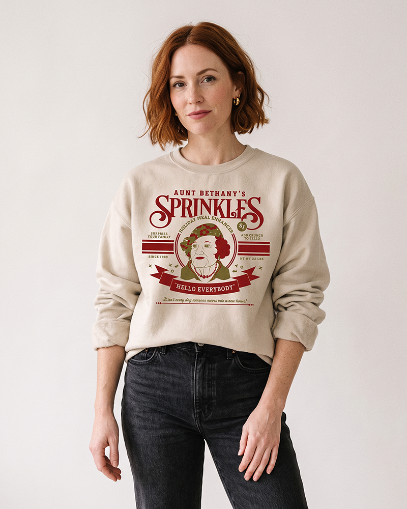 Woman wearing sand Aunt Bethany Sprinkles Christmas sweatshirt with black jeans