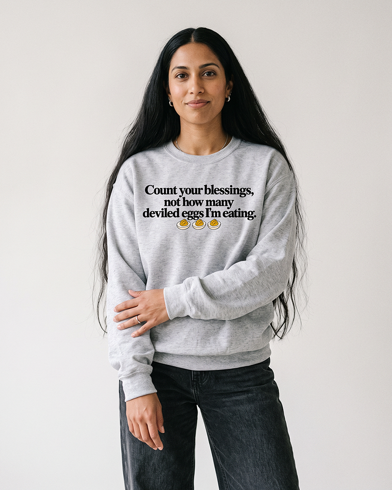 Black-haired woman modeling an ash gray Count Your Blessings Thanksgiving crewneck sweatshirt with vintage black jeans.