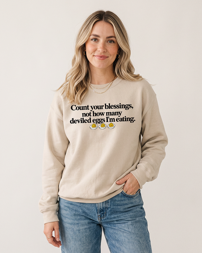 Blonde woman wearing a sand Count Your Blessings deviled eggs Thanksgiving sweatshirt with blue denim jeans.