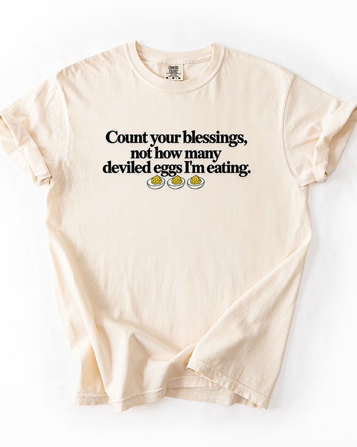 Ivory heavyweight Thanksgiving graphic tee with Count Your Blessings Not How Many Deviled Eggs I'm Eating saying.