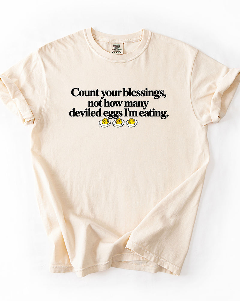 Ivory heavyweight Thanksgiving graphic tee with Count Your Blessings Not How Many Deviled Eggs I'm Eating saying.