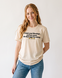 Red-haired woman wearing a natural Count Your Blessings deviled eggs Thanksgiving graphic tee with blue denim jeans.