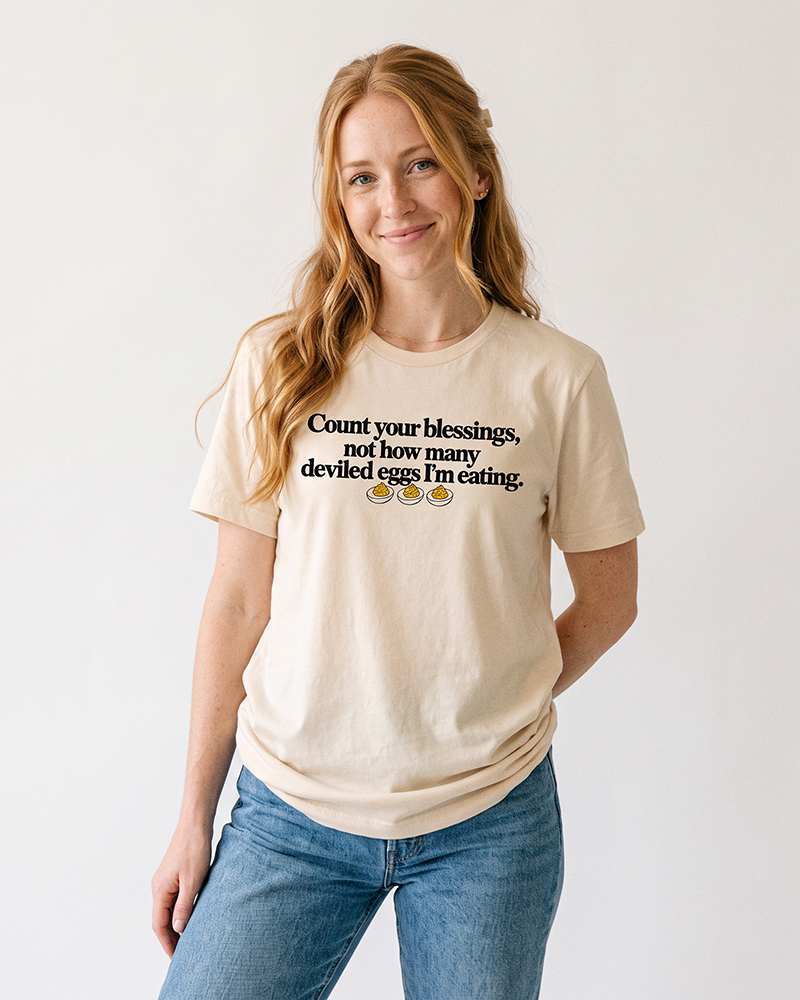 Red-haired woman wearing a natural Count Your Blessings deviled eggs Thanksgiving graphic tee with blue denim jeans.
