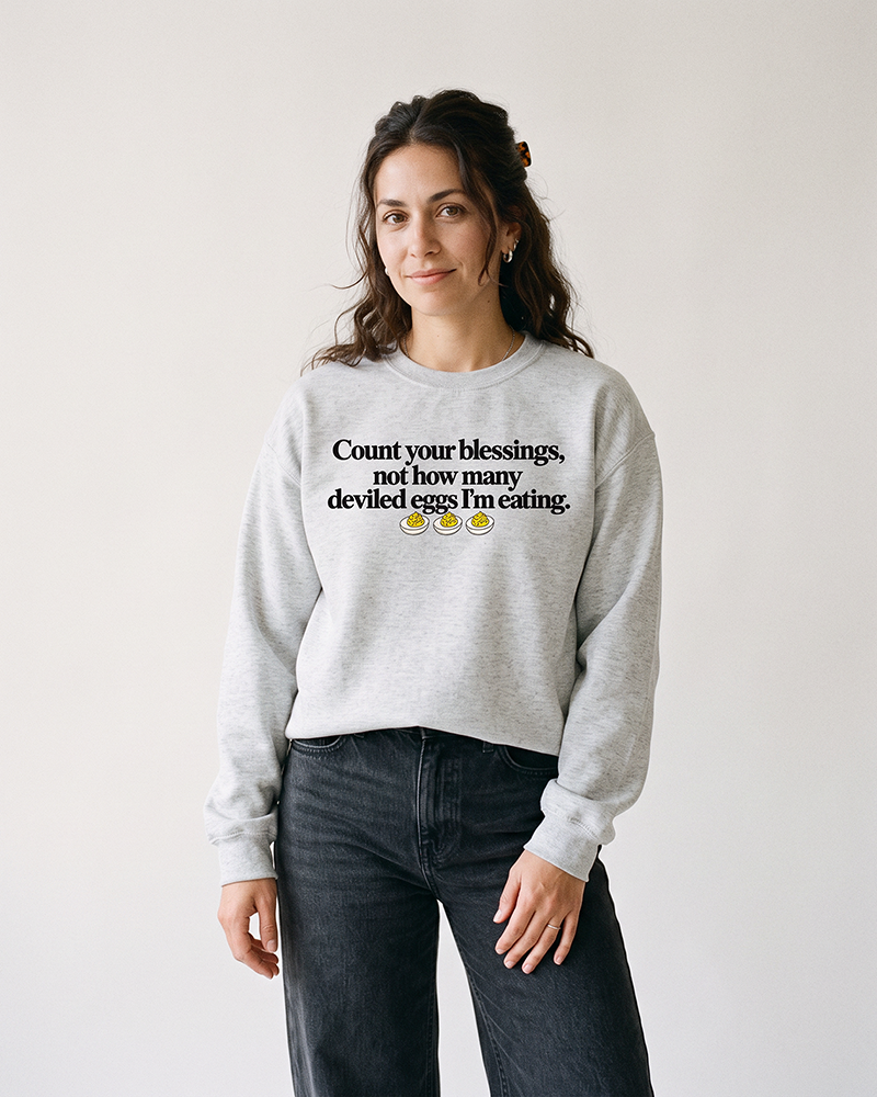 Brunette woman wearing an ash gray funny Thanksgiving deviled eggs sweatshirt with black denim jeans.