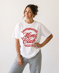 Woman wearing white Caffeinated Moms Club heavyweight graphic tee with red lettering and gray sweatpants