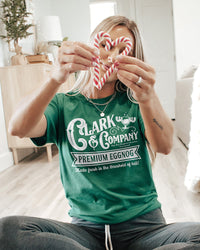 Woman wearing a green Clark & Company Christmas tee while holding candy canes during a holiday celebration.