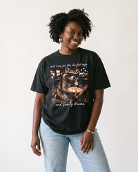 Woman wearing a Black heavyweight Thanksgiving tee featuring a sunglasses-wearing raccoon holding deviled eggs and white script lettering.