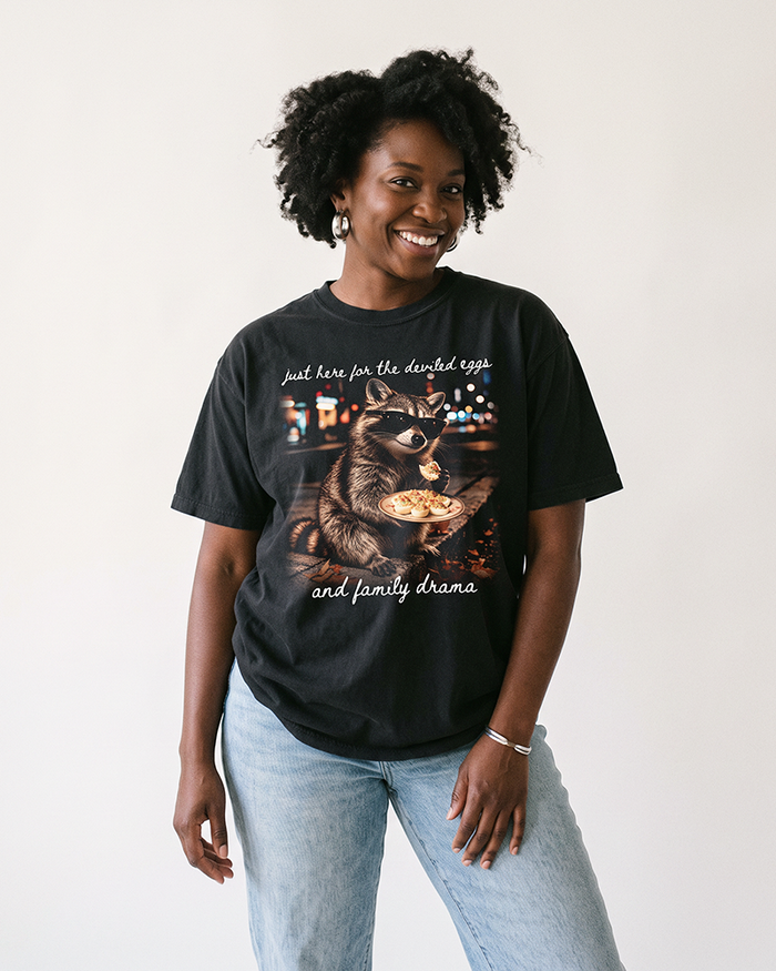 Woman wearing a Black heavyweight Thanksgiving tee featuring a sunglasses-wearing raccoon holding deviled eggs and white script lettering.