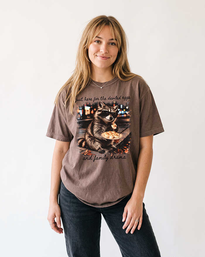 Blonde woman wearing an Espresso brown raccoon graphic tee with black Just Here for the Deviled Eggs and Family Drama lettering.