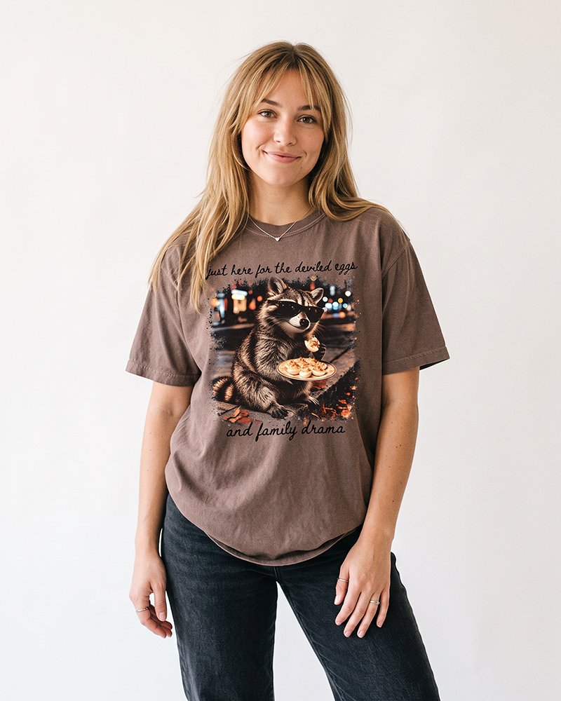 Blonde woman wearing an Espresso brown raccoon graphic tee with black Just Here for the Deviled Eggs and Family Drama lettering.