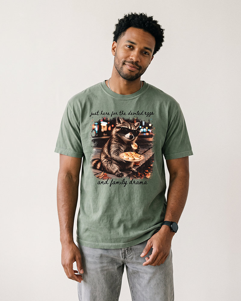 Man wearing a Hemp green heavyweight Thanksgiving graphic tee featuring a raccoon in sunglasses eating deviled eggs.