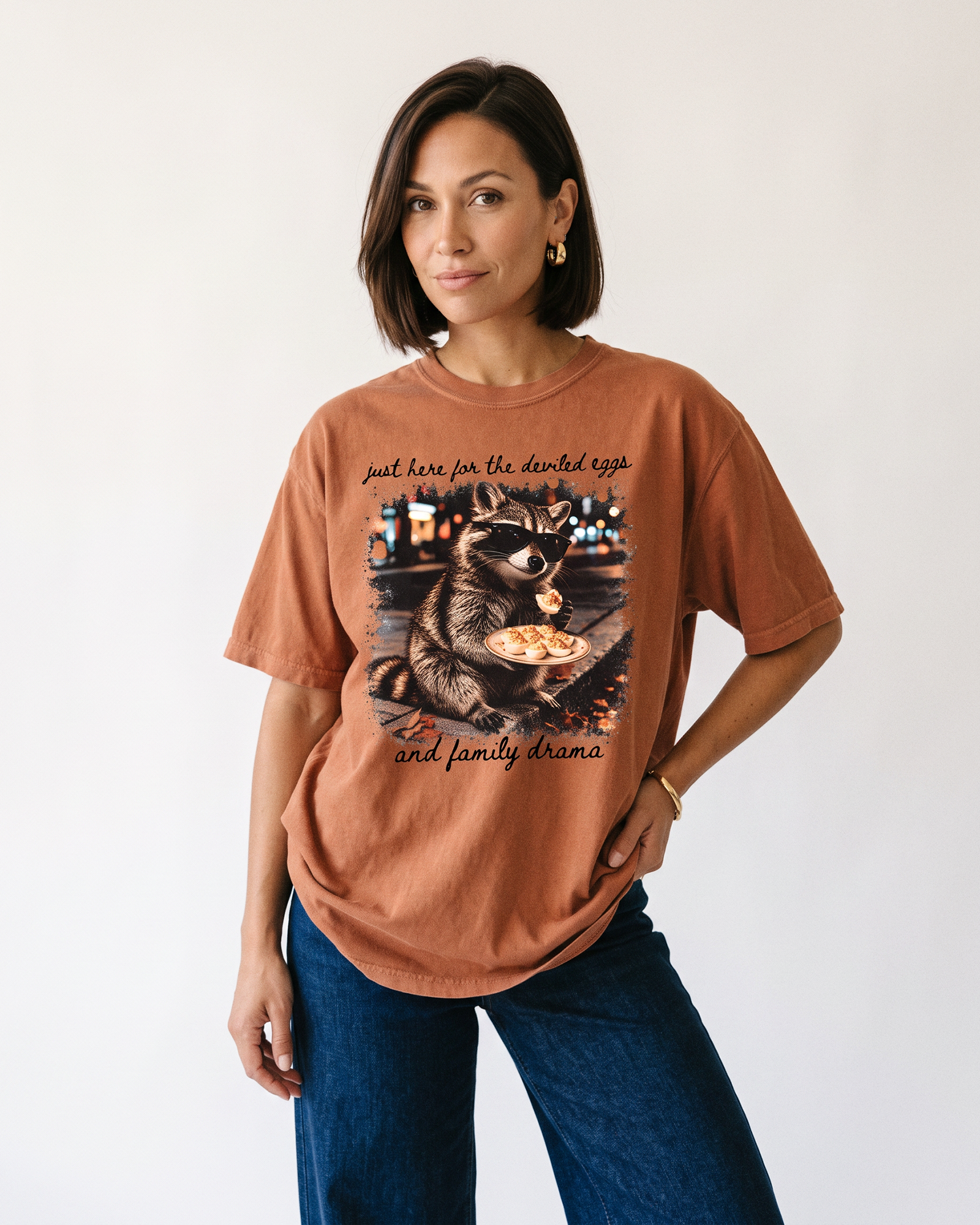 Woman wearing a Yam orange raccoon Thanksgiving graphic tee with black lettering and dark blue jeans.