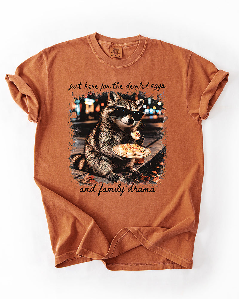 Yam orange heavyweight T-shirt featuring a raccoon wearing sunglasses, holding deviled eggs, and black family drama lettering.