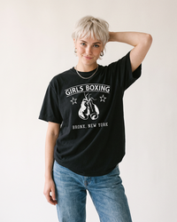 Woman with short platinum blonde hair wearing a black Girls Boxing heavyweight tee with blue jeans.