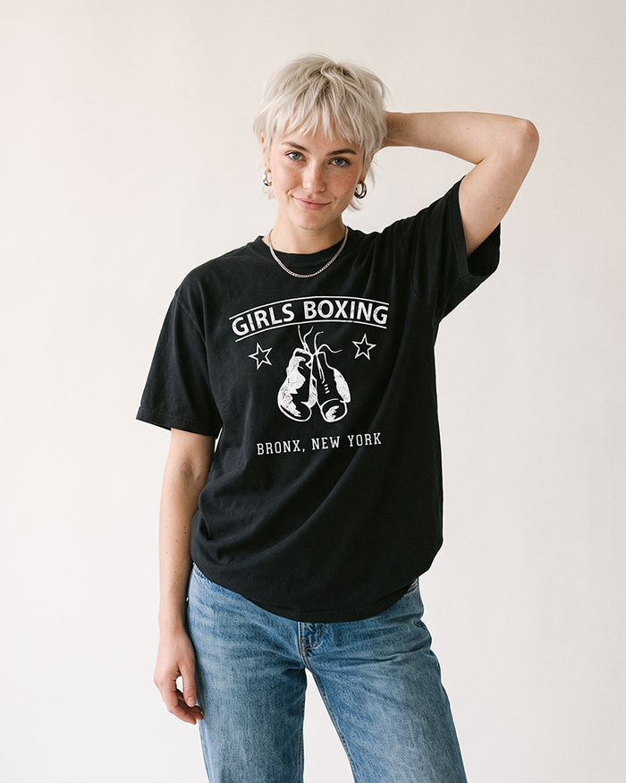 Woman with short platinum blonde hair wearing a black Girls Boxing heavyweight tee with blue jeans.