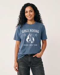Woman with long dark curly hair wearing a Denim blue Girls Boxing heavyweight t-shirt with dark jeans.