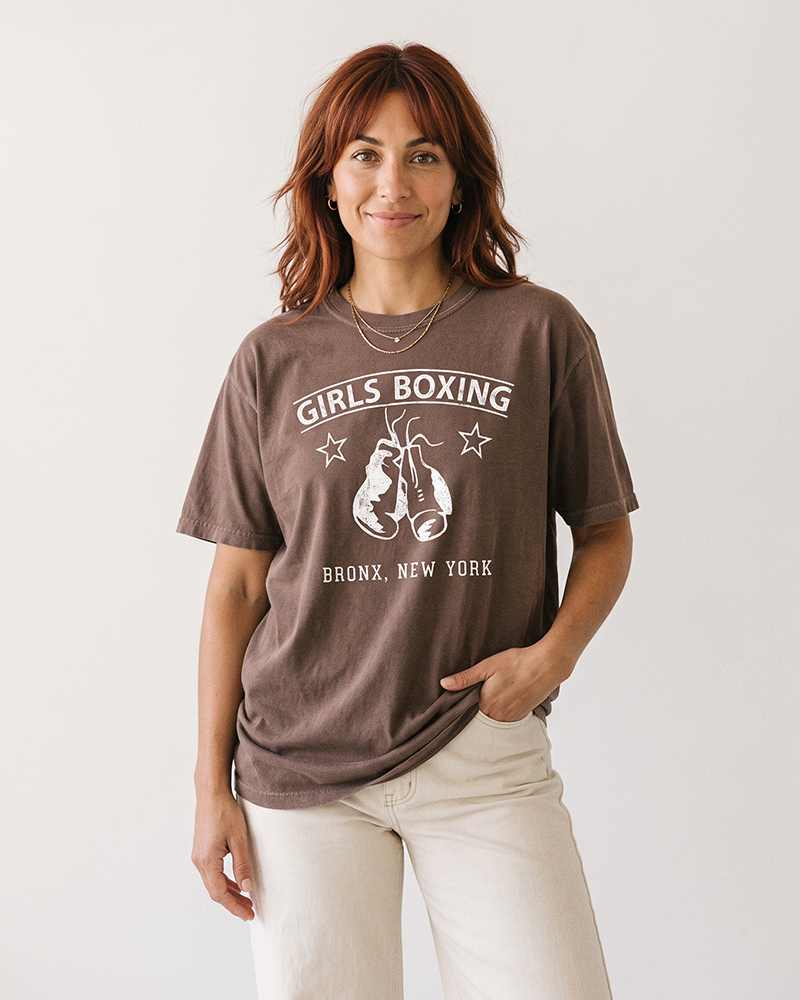 Red-haired woman wearing an Espresso brown Girls Boxing graphic tee with cream pants.