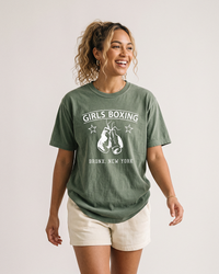 Woman with curly blonde ponytail wearing a Hemp green Girls Boxing heavyweight t-shirt with cream shorts.